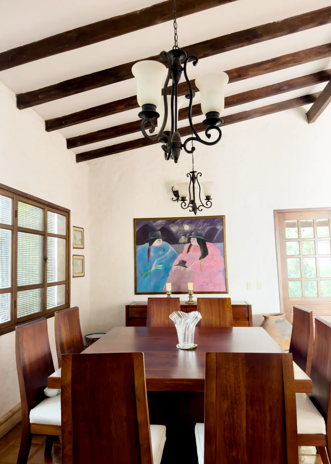 Dining table beneath wrought-iron chandeliers and a colourful painting