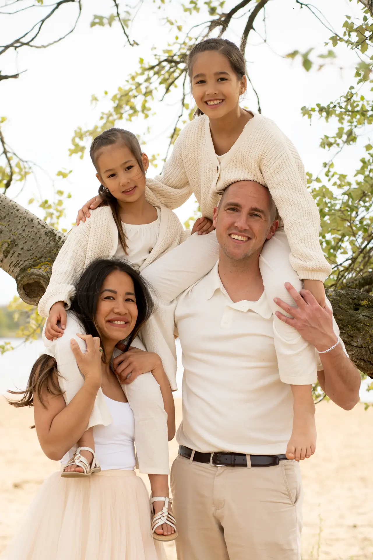 Family portrait beneath a leaning tree, daughters on their parents' shoulders