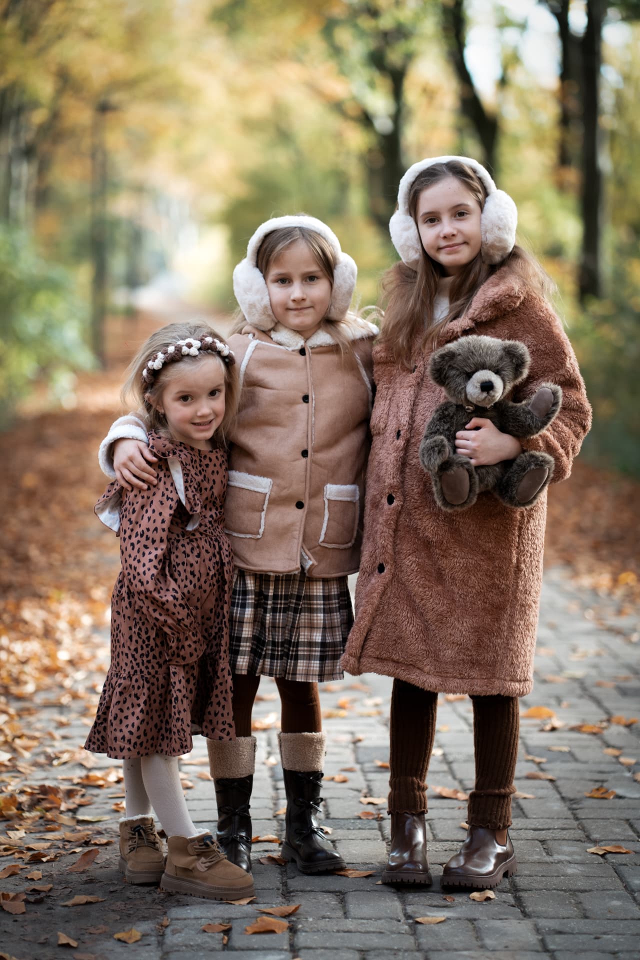 Three sisters with their teddy bear on a leaf-strewn path