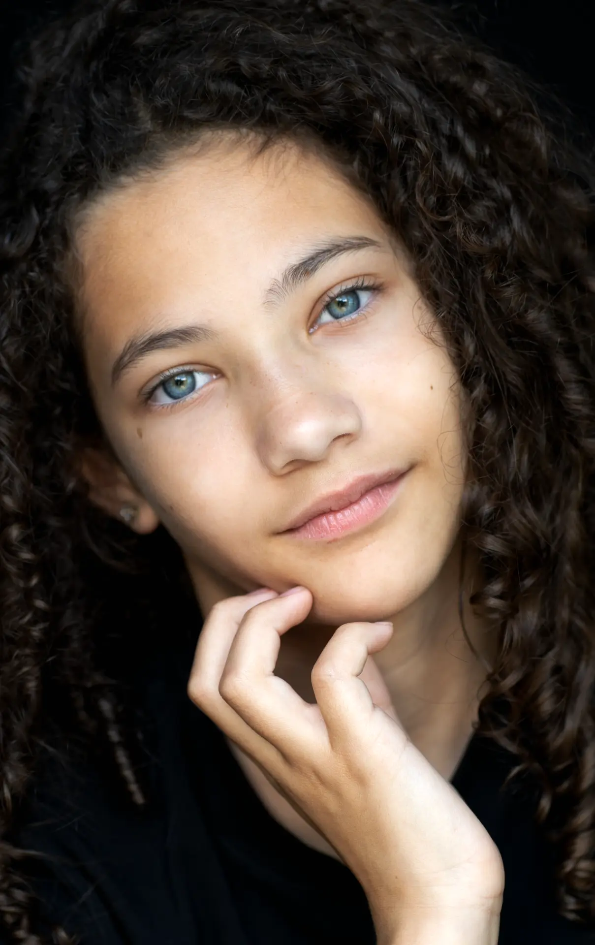 Girl with curly hair resting her hand near her chin against a black backdrop