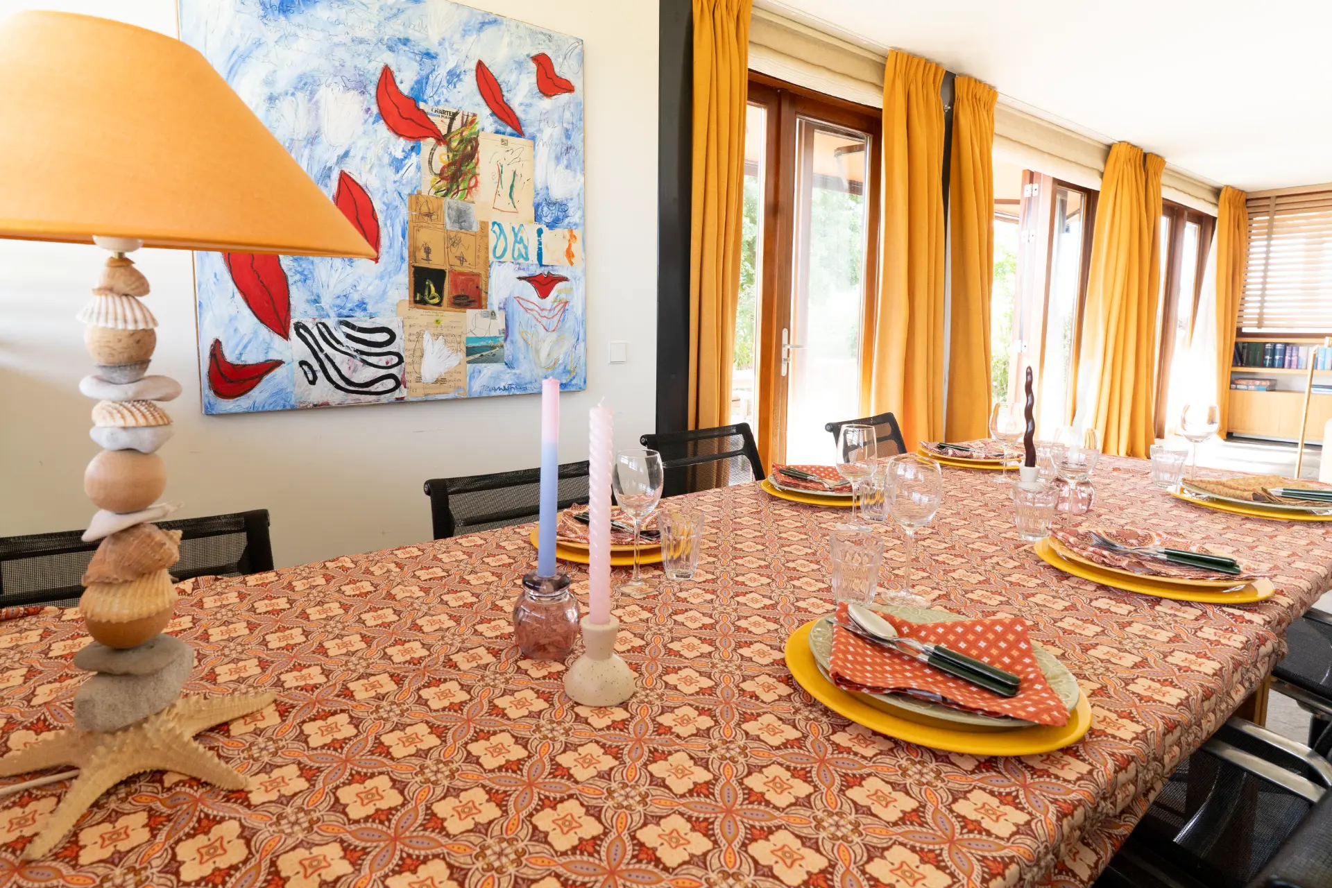 The dining table set for dinner beneath a blue painting, a lamp of stacked shells beside
