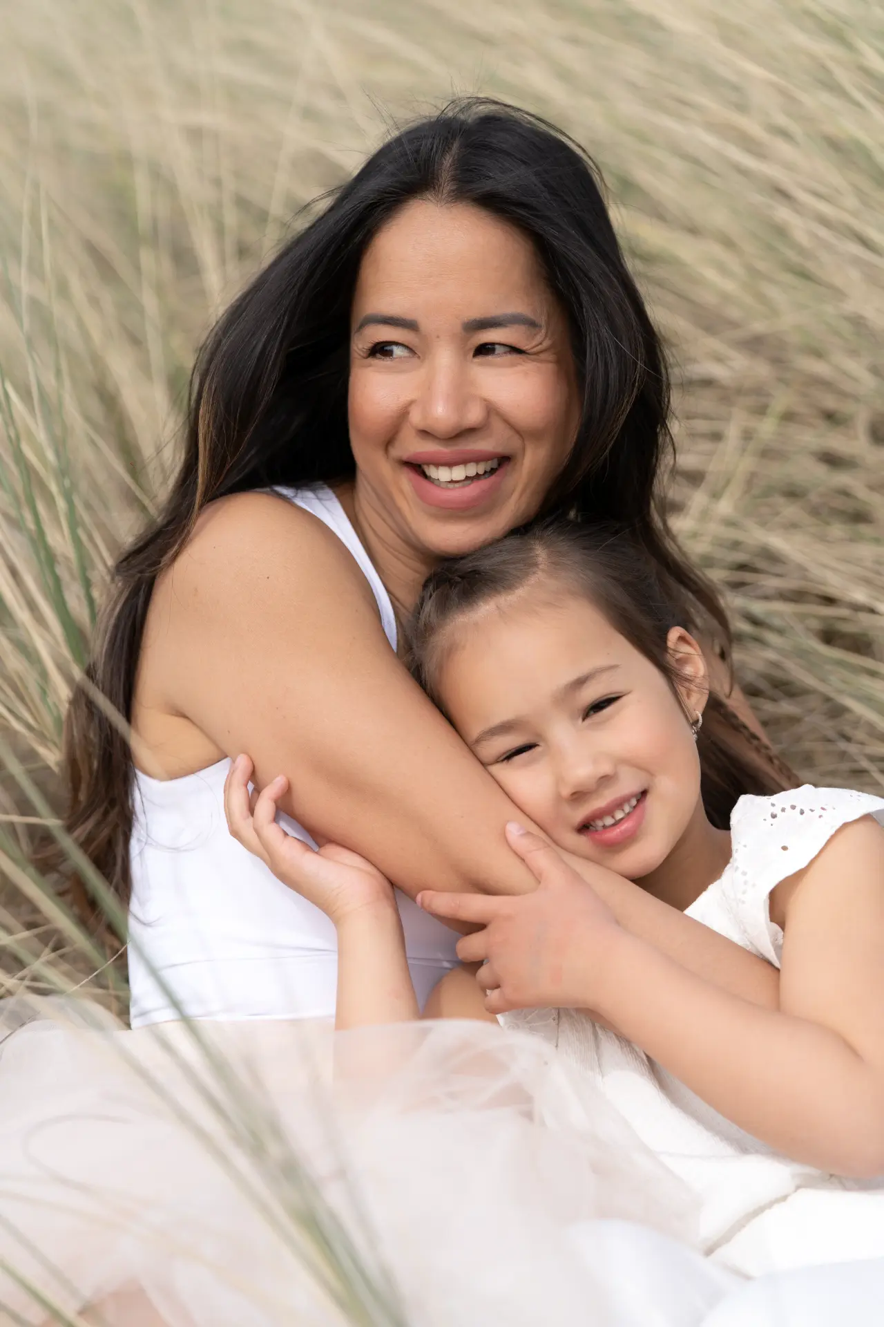 Mother laughing while her daughter hugs her arm in the dune grass