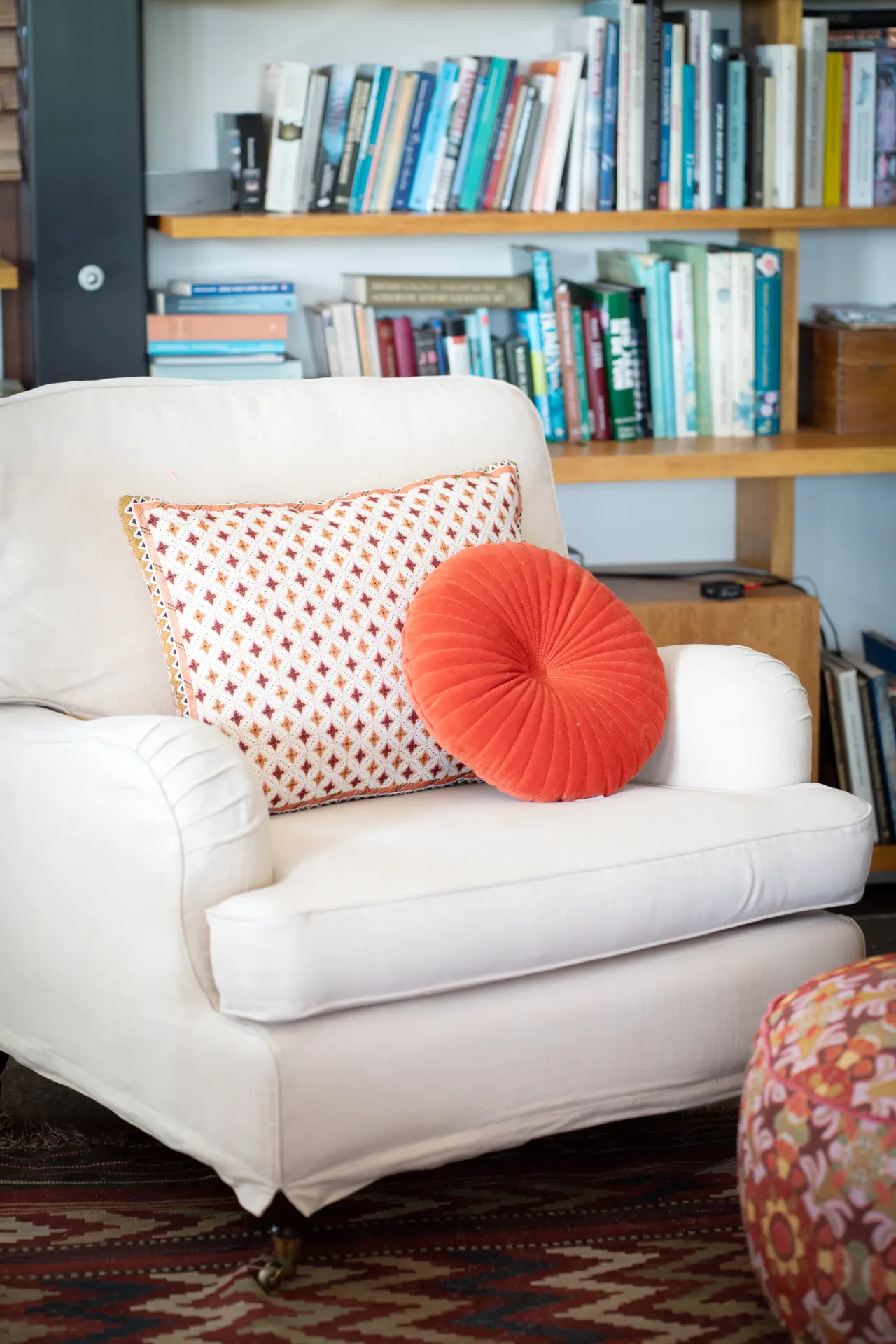 A white armchair with orange cushions in front of a wooden bookshelf