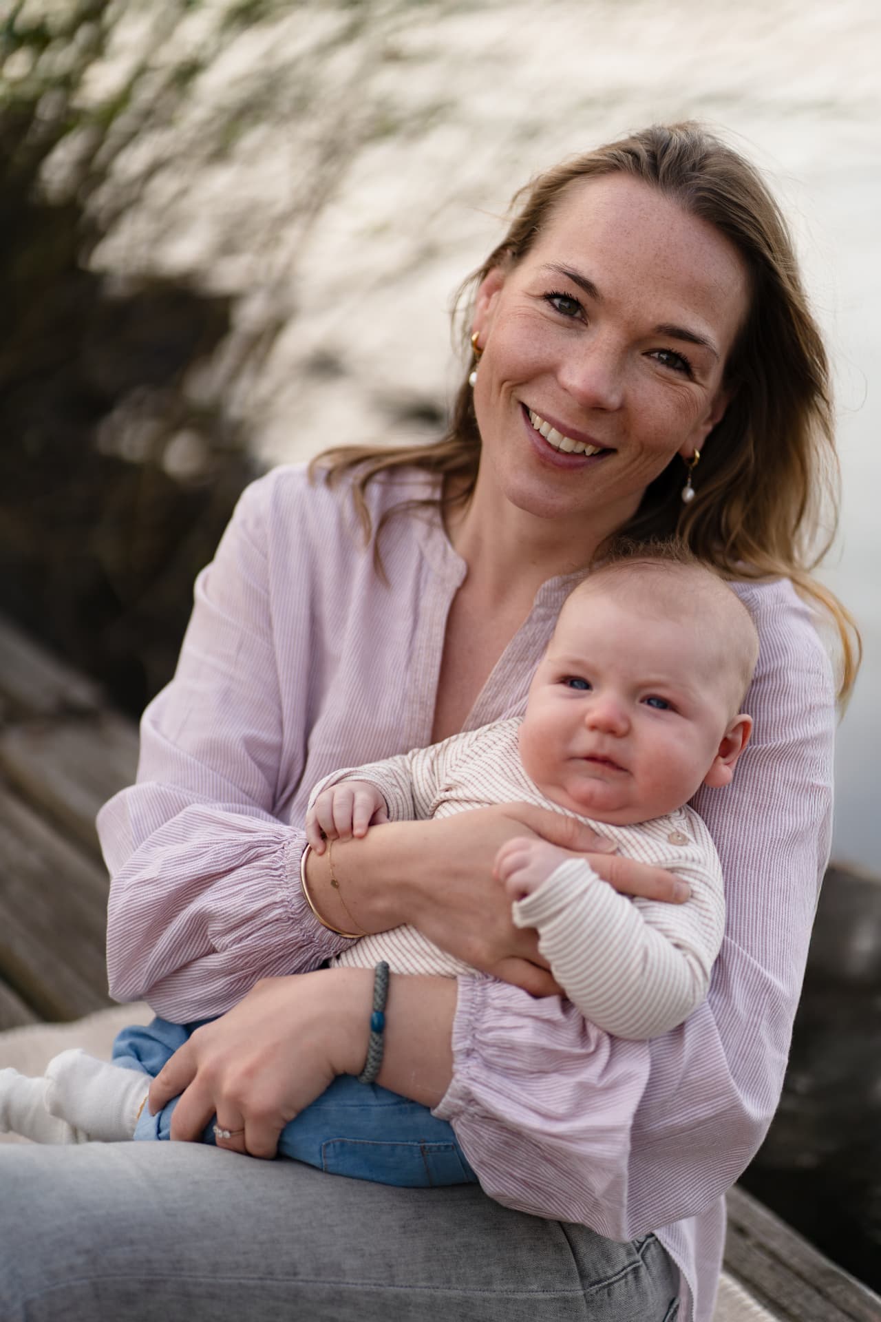 Mother smiling with her baby on her lap on a wooden jetty by the water