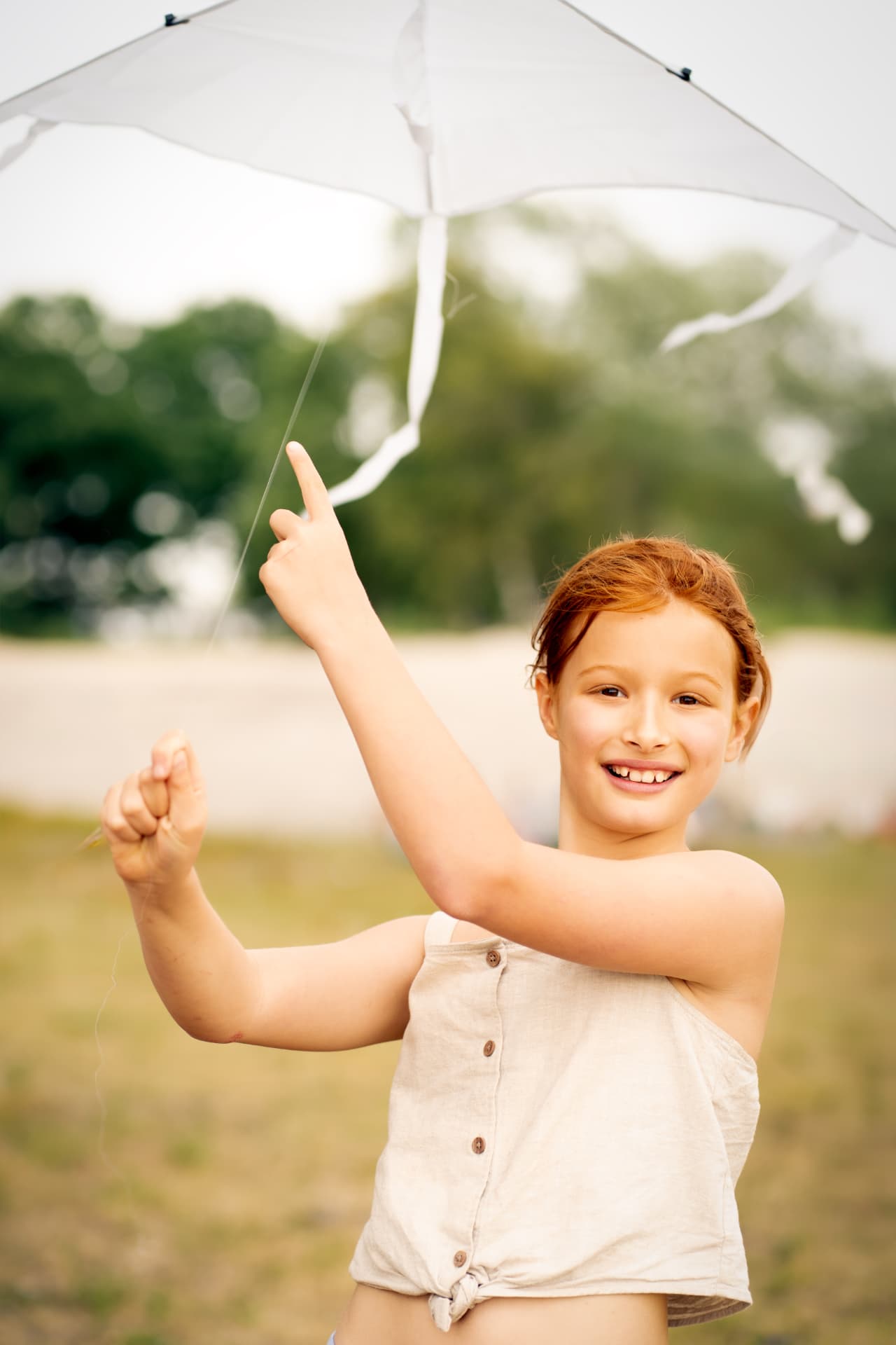 Red-haired girl in a beige top holding the string of a white kite outdoors